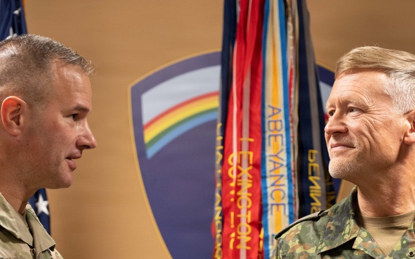 Two military officers are engaged in what appears to be a formal ceremony or award presentation. The officer on the left, identified by his name tape as 'CARPENTER,' is a U.S. Army soldier presenting or pinning something to the officer on the right, identified as 'FRANOSCH,' who is wearing a German Bundeswehr uniform. They are standing in front of military flags and a unit crest backdrop.