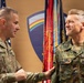 Two military officers are engaged in what appears to be a formal ceremony or award presentation. The officer on the left, identified by his name tape as 'CARPENTER,' is a U.S. Army soldier presenting or pinning something to the officer on the right, identified as 'FRANOSCH,' who is wearing a German Bundeswehr uniform. They are standing in front of military flags and a unit crest backdrop.