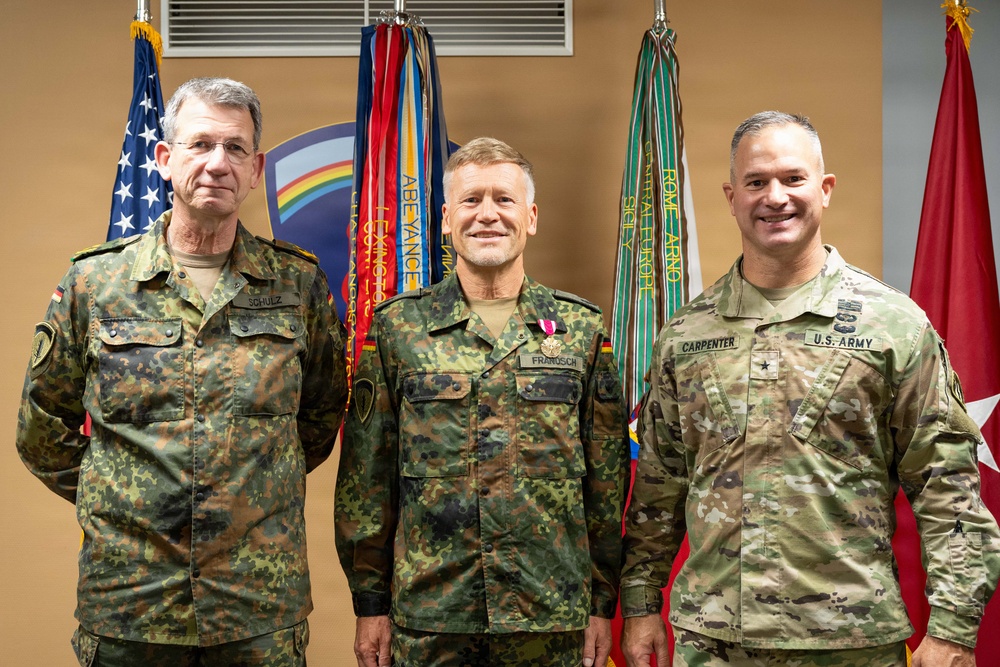 Three military officers pose for a formal photograph in front of a display of military flags and unit colors. Two officers on the left wear German Bundeswehr camouflage uniforms, while the officer on the right wears a U.S. Army OCP (Operational Camouflage Pattern) uniform. The center figure appears to be receiving or has received a medal, suggesting this is a military awards or recognition ceremony.