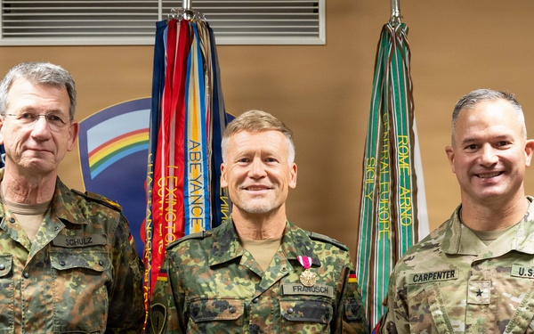 Three military officers pose for a formal photograph in front of a display of military flags and unit colors. Two officers on the left wear German Bundeswehr camouflage uniforms, while the officer on the right wears a U.S. Army OCP (Operational Camouflage Pattern) uniform. The center figure appears to be receiving or has received a medal, suggesting this is a military awards or recognition ceremony.