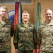 Three military officers pose for a formal photograph in front of a display of military flags and unit colors. Two officers on the left wear German Bundeswehr camouflage uniforms, while the officer on the right wears a U.S. Army OCP (Operational Camouflage Pattern) uniform. The center figure appears to be receiving or has received a medal, suggesting this is a military awards or recognition ceremony.