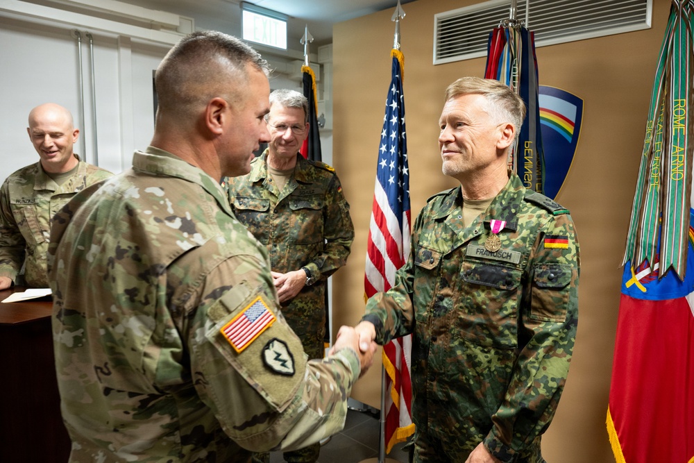 A formal military ceremony showing a US Army soldier shaking hands with a German Bundeswehr soldier named 'FRANSSCH' who is wearing a medal/decoration on his uniform. The setting appears to be an official military office or ceremony room with US and German flags visible in the background. Two additional military personnel observe the handshake from behind.