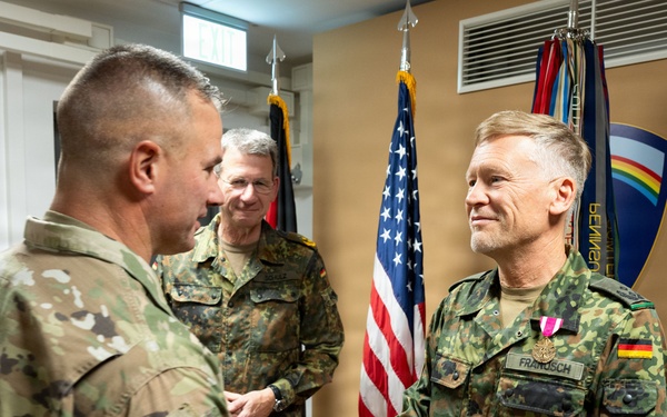 A formal military ceremony showing a US Army soldier shaking hands with a German Bundeswehr soldier named 'FRANSSCH' who is wearing a medal/decoration on his uniform. The setting appears to be an official military office or ceremony room with US and German flags visible in the background. Two additional military personnel observe the handshake from behind.