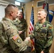 A formal military ceremony showing a US Army soldier shaking hands with a German Bundeswehr soldier named 'FRANSSCH' who is wearing a medal/decoration on his uniform. The setting appears to be an official military office or ceremony room with US and German flags visible in the background. Two additional military personnel observe the handshake from behind.
