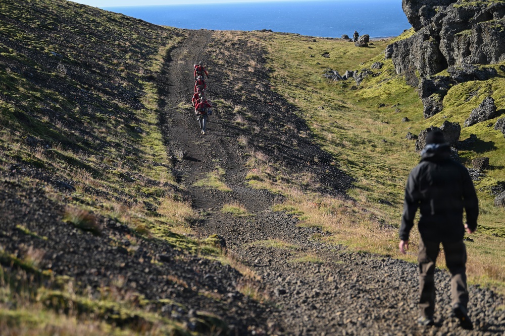 A group of hikers are ascending a steep, dark gravel trail on a rugged hillside with moss-covered volcanic rock formations on either side. In the foreground, a lone hiker dressed in black with a backpack follows behind a group of hikers in red further up the path. A body of water, likely an ocean or large lake, is visible in the background under a clear blue sky.