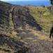 A group of hikers are ascending a steep, dark gravel trail on a rugged hillside with moss-covered volcanic rock formations on either side. In the foreground, a lone hiker dressed in black with a backpack follows behind a group of hikers in red further up the path. A body of water, likely an ocean or large lake, is visible in the background under a clear blue sky.