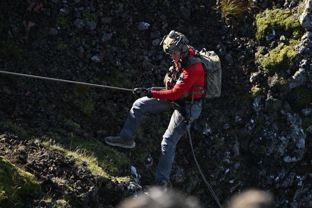A person is rappelling or abseiling down a rocky cliff face while holding a rope. The individual is equipped with tactical/military-style gear including a helmet, backpack, harness, and gloves. The rocky terrain features patches of moss and grass, suggesting a rugged outdoor environment.