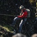 A person is rappelling or abseiling down a rocky cliff face while holding a rope. The individual is equipped with tactical/military-style gear including a helmet, backpack, harness, and gloves. The rocky terrain features patches of moss and grass, suggesting a rugged outdoor environment.