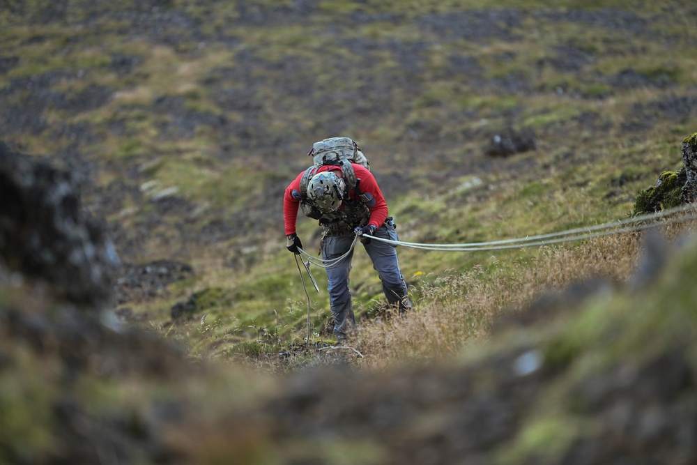 A person equipped with climbing gear, a helmet, and a large backpack is navigating a steep grassy hillside while managing a rope. The terrain appears to be a rugged, moss-covered highland or mountainous landscape, likely in a northern or subarctic region. The individual appears to be engaged in a technical outdoor activity such as mountaineering, search and rescue, or military training.