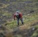 A person equipped with climbing gear, a helmet, and a large backpack is navigating a steep grassy hillside while managing a rope. The terrain appears to be a rugged, moss-covered highland or mountainous landscape, likely in a northern or subarctic region. The individual appears to be engaged in a technical outdoor activity such as mountaineering, search and rescue, or military training.