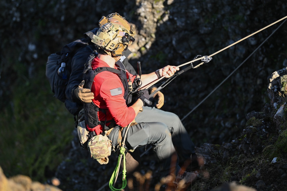 A military or search-and-rescue operation is underway on a rocky cliff face. A helmeted operator wearing multicam gear and night-vision mount is securing or extracting a civilian wearing a red long-sleeve shirt with a US flag patch. Both individuals are suspended on ropes/rigging attached to a carabiner system above them.