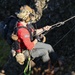 A military or search-and-rescue operation is underway on a rocky cliff face. A helmeted operator wearing multicam gear and night-vision mount is securing or extracting a civilian wearing a red long-sleeve shirt with a US flag patch. Both individuals are suspended on ropes/rigging attached to a carabiner system above them.