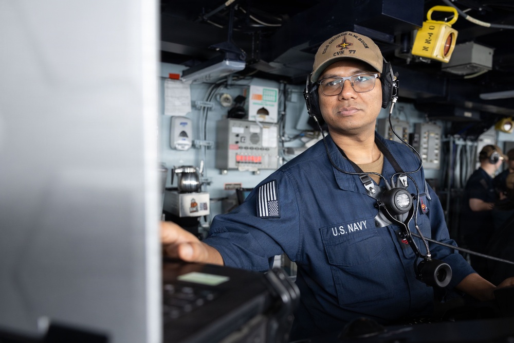 A U.S. Navy sailor is stationed at a control console aboard USS George H.W. Bush (CVN-77), wearing standard Navy working uniform with safety glasses and a communications headset. The sailor is positioned at what appears to be a bridge or combat information center workstation. The background shows various naval control panels, switches, and equipment typical of a warship's operational spaces.
