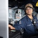 A U.S. Navy sailor is stationed at a control console aboard USS George H.W. Bush (CVN-77), wearing standard Navy working uniform with safety glasses and a communications headset. The sailor is positioned at what appears to be a bridge or combat information center workstation. The background shows various naval control panels, switches, and equipment typical of a warship's operational spaces.