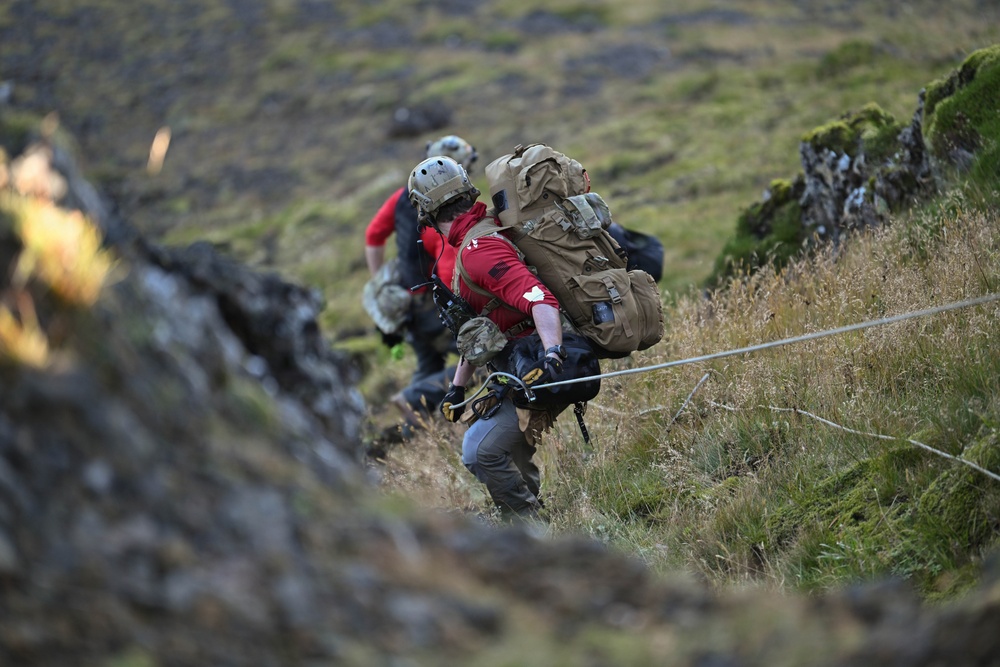 Two individuals are ascending a steep, grassy and rocky hillside while holding onto a guide rope. The person in the foreground wears a red jacket, tactical helmet, and carries a large tan/coyote-colored military-style backpack with climbing gear attached. The terrain appears rugged with moss-covered rocks and dry grass, suggesting a mountainous or highland environment.