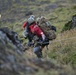 Two individuals are ascending a steep, grassy and rocky hillside while holding onto a guide rope. The person in the foreground wears a red jacket, tactical helmet, and carries a large tan/coyote-colored military-style backpack with climbing gear attached. The terrain appears rugged with moss-covered rocks and dry grass, suggesting a mountainous or highland environment.