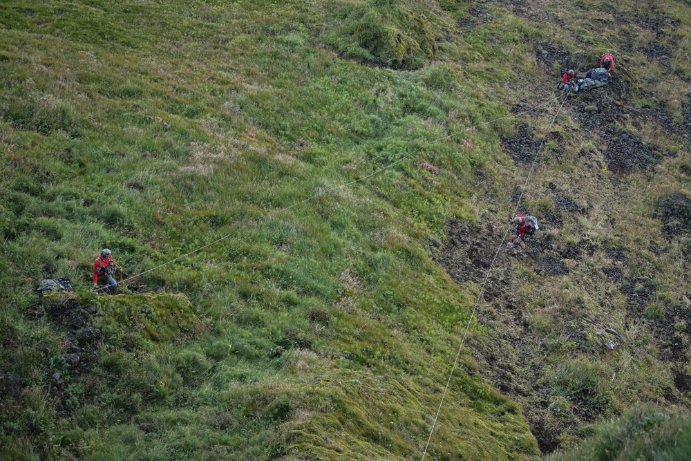 The image shows a steep, grassy and rocky hillside or cliff face with multiple individuals engaged in a rope-assisted descent or rescue operation. At least four people are visible at different elevations on the slope, connected by ropes. The terrain is rugged, featuring patches of dark volcanic rock interspersed with dense green vegetation.