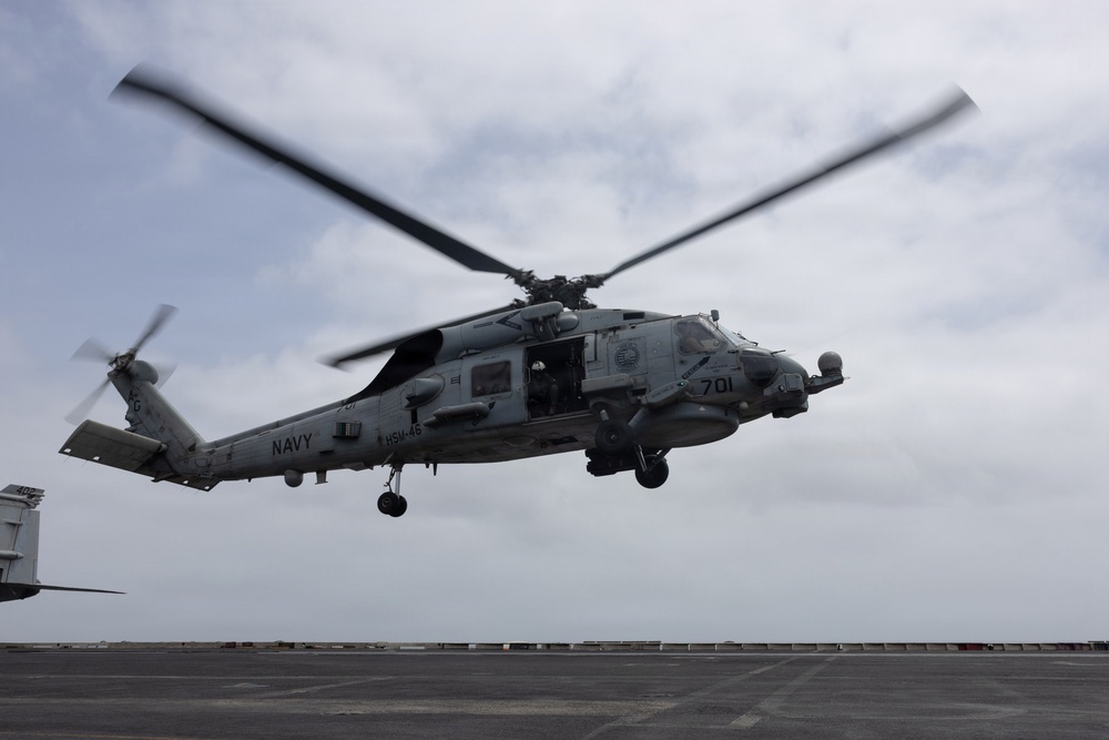 A U.S. Navy MH-60R Seahawk helicopter marked '701' and assigned to HSM-46 is hovering low over what appears to be the flight deck of a naval vessel. The helicopter's main rotor blades are in motion, visible as a blur, indicating active flight operations. The image was taken from a low angle on the flight deck, with an overcast sky in the background.