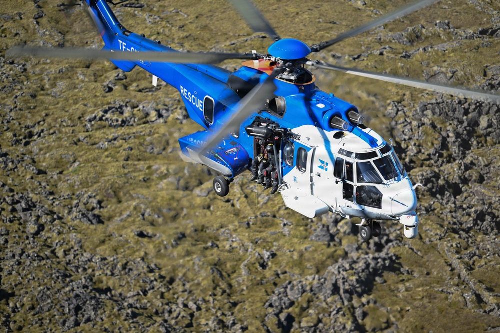 A blue and white search and rescue helicopter is captured in flight over a rocky, moss-covered terrain. The aircraft is clearly marked 'RESCUE' on its fuselage and bears a registration beginning with 'TF-', suggesting Icelandic registration. The photograph is taken from an aerial perspective, likely from another aircraft, providing a detailed view of the helicopter's top and front sections.