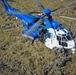 A blue and white search and rescue helicopter is captured in flight over a rocky, moss-covered terrain. The aircraft is clearly marked 'RESCUE' on its fuselage and bears a registration beginning with 'TF-', suggesting Icelandic registration. The photograph is taken from an aerial perspective, likely from another aircraft, providing a detailed view of the helicopter's top and front sections.