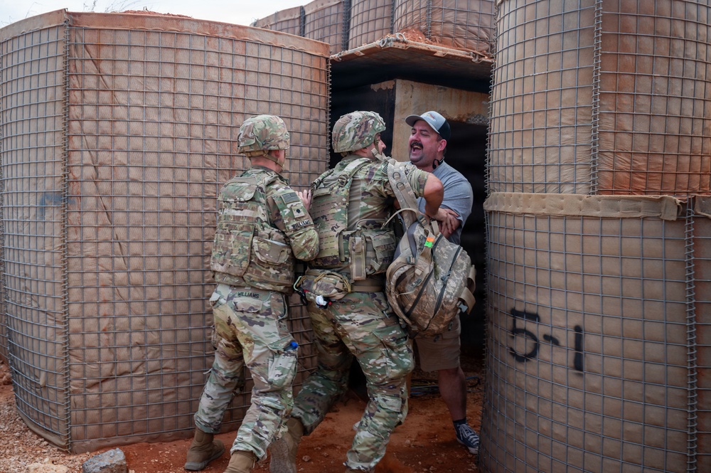 Two U.S. Army soldiers in MultiCam/OCP camouflage uniforms are engaged with a civilian male in a gray t-shirt and baseball cap near what appear to be HESCO barrier walls. The interaction appears to be a training or role-player scenario, likely at a military training facility, given the structured environment and the civilian's apparent role-player positioning. The barriers are labeled '5-1', suggesting a designated training lane or position.