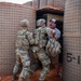 Two U.S. Army soldiers in MultiCam/OCP camouflage uniforms are engaged with a civilian male in a gray t-shirt and baseball cap near what appear to be HESCO barrier walls. The interaction appears to be a training or role-player scenario, likely at a military training facility, given the structured environment and the civilian's apparent role-player positioning. The barriers are labeled '5-1', suggesting a designated training lane or position.
