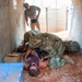 The image shows what appears to be a military or tactical training exercise taking place inside a concrete structure. Two individuals in military camouflage uniforms are attending to a person lying on the ground, while a third person in civilian clothing observes from behind. The setting includes props such as cardboard boxes and a blue container, consistent with a simulated scenario environment.
