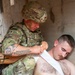 A U.S. Army soldier identified by name tape as 'Williams' is applying a bandage or dressing to a shirtless casualty leaning against a concrete wall. The soldier is wearing full combat gear including a multicam helmet and body armor. This appears to be a combat casualty care scenario, either a training exercise or an actual field medical treatment situation.