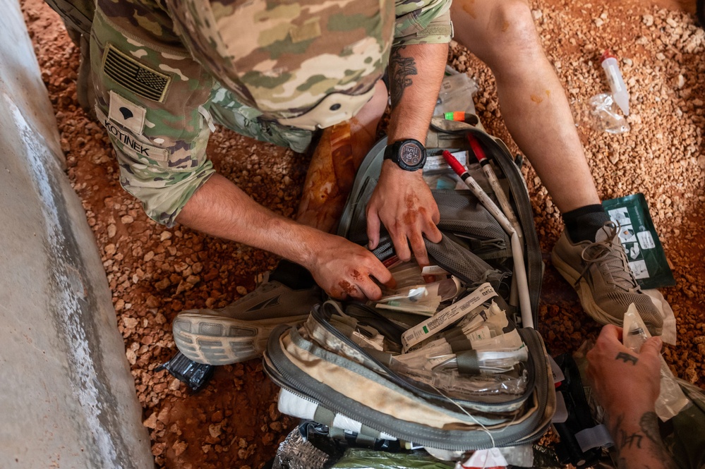 A U.S. Army soldier identified by the name tape 'KOTINEK' is kneeling on gravel terrain and accessing a medical kit or tactical bag filled with packaged medical supplies. The soldier is wearing MultiCam (OCP) camouflage uniform with a U.S. flag patch and appears to have blood or iodine-stained hands. A second individual in civilian-style clothing with tattoos is visible nearby, also handling medical supplies.