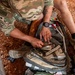 A U.S. Army soldier identified by the name tape 'KOTINEK' is kneeling on gravel terrain and accessing a medical kit or tactical bag filled with packaged medical supplies. The soldier is wearing MultiCam (OCP) camouflage uniform with a U.S. flag patch and appears to have blood or iodine-stained hands. A second individual in civilian-style clothing with tattoos is visible nearby, also handling medical supplies.