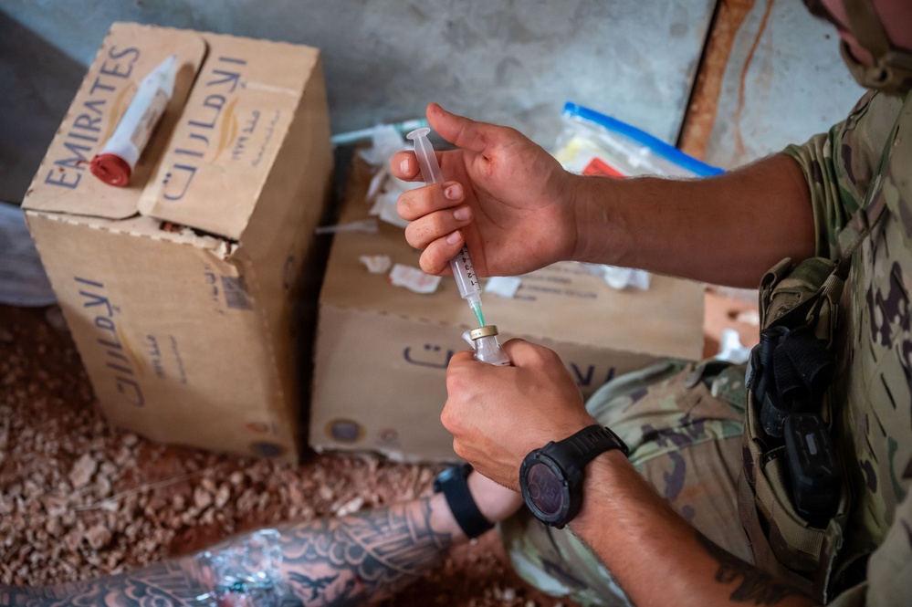 A person wearing military camouflage clothing is drawing medication from a vial into a syringe, consistent with medical preparation activities. The individual has visible tattoos on their forearm and wears a tactical watch. Cardboard boxes with Arabic script and 'EMIRATES' branding are visible in the background, suggesting a field medical or humanitarian aid context.