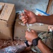 A person wearing military camouflage clothing is drawing medication from a vial into a syringe, consistent with medical preparation activities. The individual has visible tattoos on their forearm and wears a tactical watch. Cardboard boxes with Arabic script and 'EMIRATES' branding are visible in the background, suggesting a field medical or humanitarian aid context.