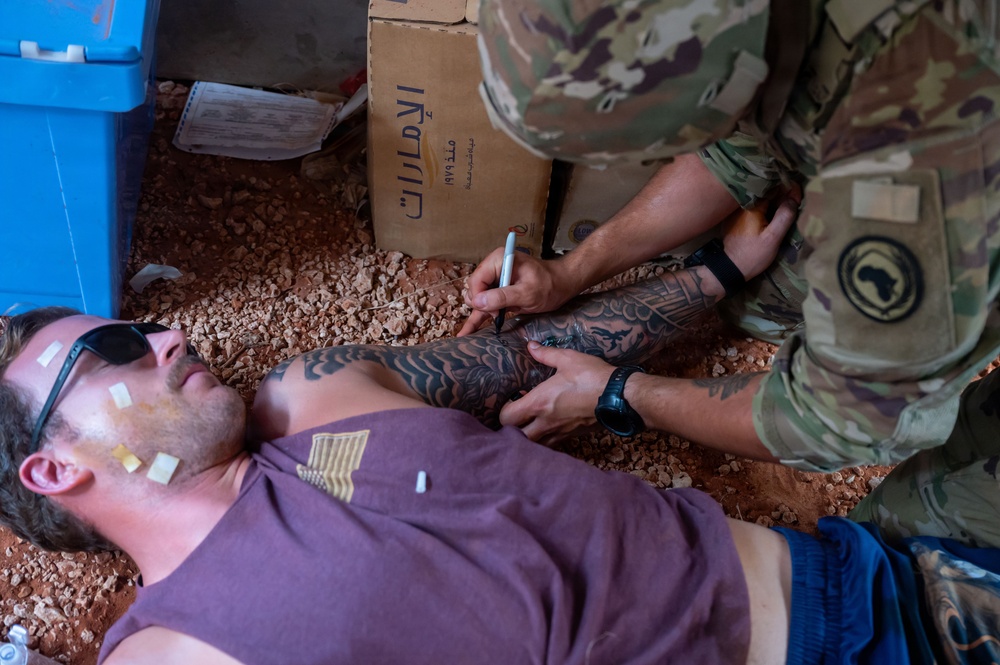 A person in military camouflage uniform is attending to a casualty lying on the ground, appearing to write on the person's arm with a marker. The casualty has simulated wound markers (moulage stickers) applied to their face and torso, suggesting this is a medical training exercise. A cardboard box with Arabic text is visible in the background.