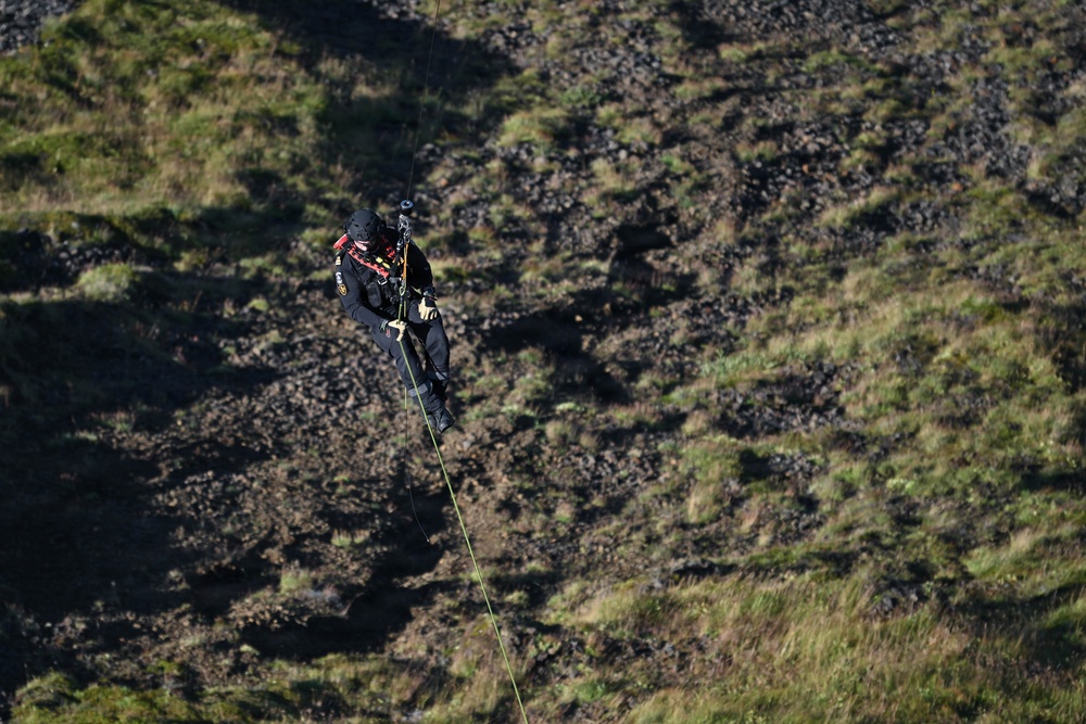 A person in full black tactical/rescue gear is suspended on a rope, rappelling or being lowered down a steep rocky hillside. The individual is equipped with a helmet, harness, and multiple attached devices, suggesting a professional rescue or technical climbing operation. The background shows a rugged terrain of dark rocks interspersed with patches of green vegetation.