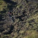A person in full black tactical/rescue gear is suspended on a rope, rappelling or being lowered down a steep rocky hillside. The individual is equipped with a helmet, harness, and multiple attached devices, suggesting a professional rescue or technical climbing operation. The background shows a rugged terrain of dark rocks interspersed with patches of green vegetation.