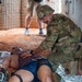 A soldier in full combat gear (multicam uniform and helmet) is providing medical assistance to a casualty lying on the ground in what appears to be a training exercise or field medical scenario. The casualty is a civilian-clothed individual lying supine on red dirt ground surrounded by medical supplies. The setting appears to be a constructed training environment based on the wire mesh walls and numbered markings.