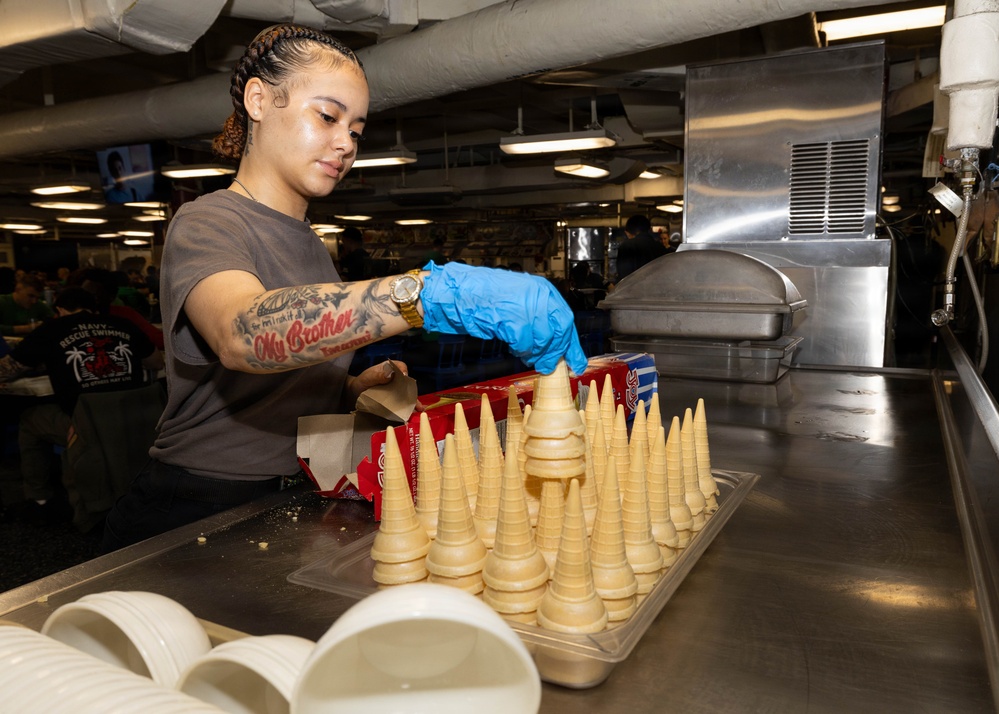 A person wearing blue nitrile gloves and a gray t-shirt is arranging ice cream cones on a stainless steel tray in what appears to be a military ship galley or large institutional kitchen. The individual has visible tattoos on their arm, including text reading 'My Brother,' and is wearing a gold watch. The background shows a busy dining facility with other personnel and industrial kitchen equipment.