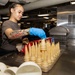 A person wearing blue nitrile gloves and a gray t-shirt is arranging ice cream cones on a stainless steel tray in what appears to be a military ship galley or large institutional kitchen. The individual has visible tattoos on their arm, including text reading 'My Brother,' and is wearing a gold watch. The background shows a busy dining facility with other personnel and industrial kitchen equipment.