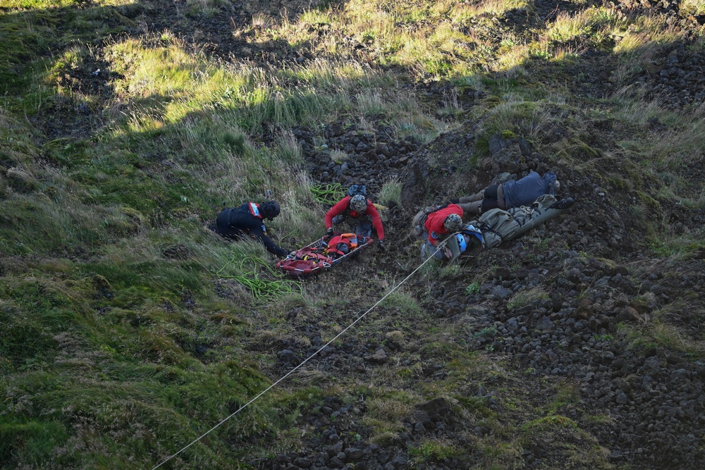 A mountain rescue operation is underway on a steep, rocky and grassy hillside. Three rescue personnel are working together around an orange rescue stretcher (litter/basket), with ropes visible for safety and lowering equipment. The terrain appears to be volcanic or rugged highland, consistent with locations such as Iceland or similar environments.