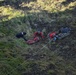 A mountain rescue operation is underway on a steep, rocky and grassy hillside. Three rescue personnel are working together around an orange rescue stretcher (litter/basket), with ropes visible for safety and lowering equipment. The terrain appears to be volcanic or rugged highland, consistent with locations such as Iceland or similar environments.