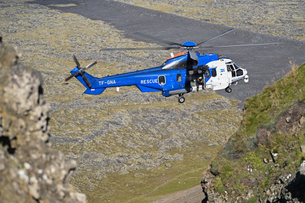 A blue and white rescue helicopter with registration TF-GNA is captured in flight over a rugged, moss-covered rocky terrain. The helicopter is clearly marked 'RESCUE' on its fuselage and appears to be operating in a mountainous or volcanic landscape, consistent with Icelandic geography. The image is taken from an elevated rocky vantage point, providing a dramatic aerial perspective.