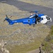 A blue and white rescue helicopter with registration TF-GNA is captured in flight over a rugged, moss-covered rocky terrain. The helicopter is clearly marked 'RESCUE' on its fuselage and appears to be operating in a mountainous or volcanic landscape, consistent with Icelandic geography. The image is taken from an elevated rocky vantage point, providing a dramatic aerial perspective.