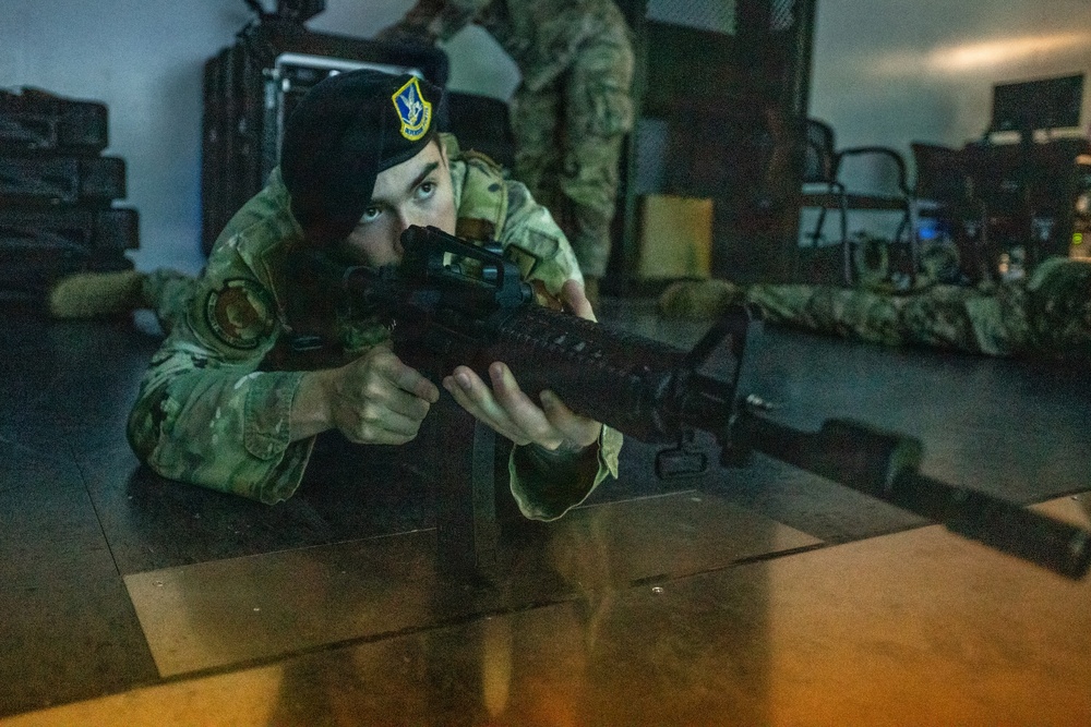 A military personnel member in camouflage uniform and black beret is in a prone firing position, aiming an M4-style carbine rifle forward. The setting appears to be an indoor training facility or exercise environment. Additional personnel in camouflage uniforms are visible in the background.