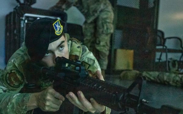 A military personnel member in camouflage uniform and black beret is in a prone firing position, aiming an M4-style carbine rifle forward. The setting appears to be an indoor training facility or exercise environment. Additional personnel in camouflage uniforms are visible in the background.