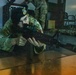 A military personnel member in camouflage uniform and black beret is in a prone firing position, aiming an M4-style carbine rifle forward. The setting appears to be an indoor training facility or exercise environment. Additional personnel in camouflage uniforms are visible in the background.