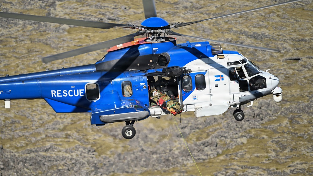 A blue and white search and rescue helicopter is captured mid-flight over rocky terrain, actively conducting a rescue operation. A person on a stretcher is being hoisted into the open side door of the helicopter via a cable. The aircraft displays the word 'RESCUE' on its fuselage and bears what appears to be an Icelandic flag emblem.