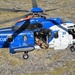 A blue and white search and rescue helicopter is captured mid-flight over rocky terrain, actively conducting a rescue operation. A person on a stretcher is being hoisted into the open side door of the helicopter via a cable. The aircraft displays the word 'RESCUE' on its fuselage and bears what appears to be an Icelandic flag emblem.