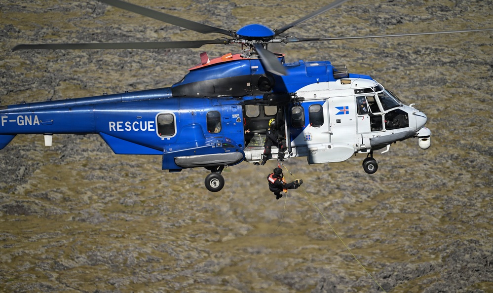 A blue and white rescue helicopter with registration 'TF-GNA' and the word 'RESCUE' painted on its fuselage is conducting a hoist operation over rocky terrain. A rescue operative is being lowered or raised on a cable beneath the aircraft, while another crew member is visible in the open side door. The Icelandic flag emblem is visible on the nose of the helicopter, indicating this is an Icelandic Coast Guard or rescue service aircraft.