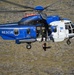 A blue and white rescue helicopter with registration 'TF-GNA' and the word 'RESCUE' painted on its fuselage is conducting a hoist operation over rocky terrain. A rescue operative is being lowered or raised on a cable beneath the aircraft, while another crew member is visible in the open side door. The Icelandic flag emblem is visible on the nose of the helicopter, indicating this is an Icelandic Coast Guard or rescue service aircraft.