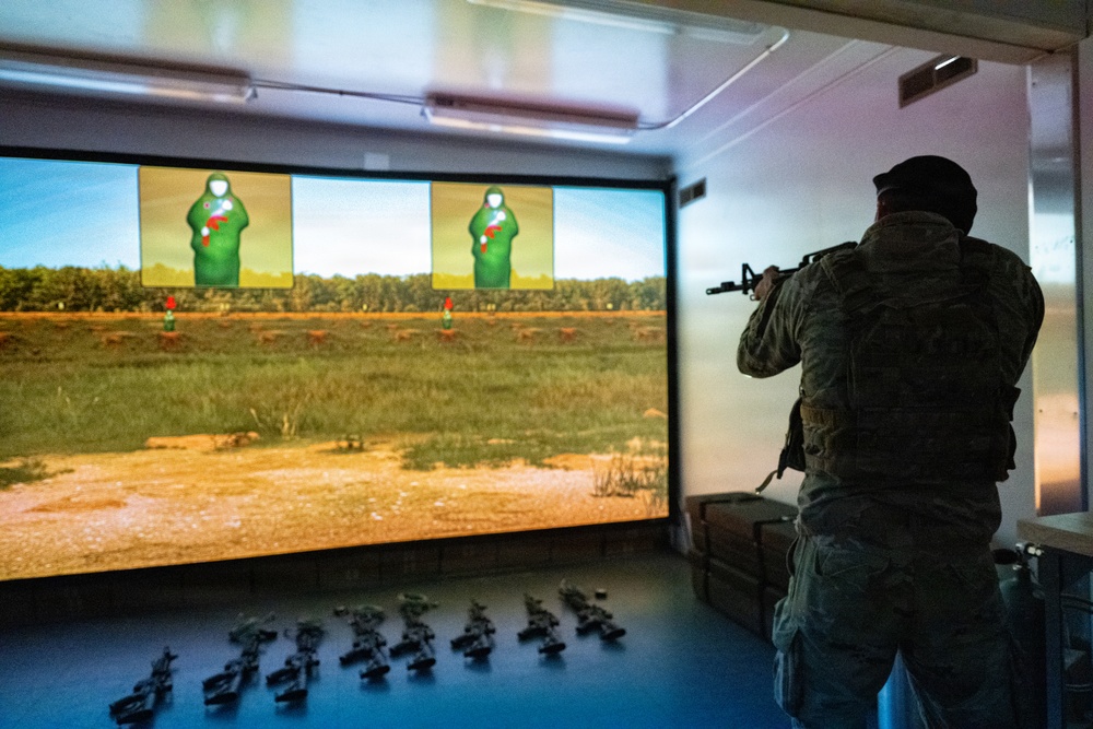 A soldier in full combat gear and camouflage uniform is using a firearms simulation training system, aiming a rifle at projected targets on a large screen. The simulation displays a realistic outdoor range environment with green humanoid silhouette targets showing red hit markers. Multiple training rifles are laid out on the floor behind the soldier, suggesting a group training session.